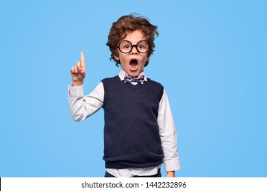 Smart little boy in round glasses opening mouth and pointing up while having idea against blue background - Shutterstock ID 1442232896