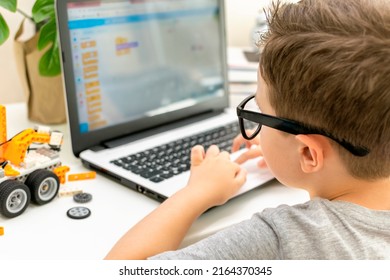 Smart little boy student in glasses builds and programs a robotic vehicle codes an electronic toy. Child using laptop in science class, his hands typing on keyboard. Programming lesson. Rear view.