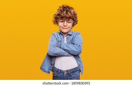 Smart little ginger nerd boy with curly hair wearing denim outfit and round glasses standing with arms crossed against yellow background and looking at camera - Powered by Shutterstock