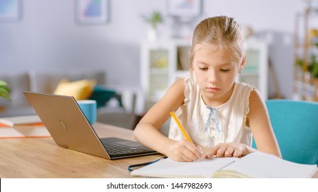 Smart Little Girl Does Homework in Her Living Room. She's Sitting at Her Desk Writes with a Pen in Her Textbooks and  Uses Laptop.