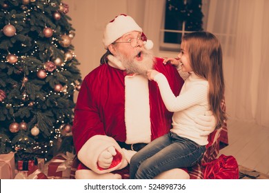 Smart Little Girl Sitting On The Lap Of Old Santa And  Pulling His Beard,  Check He Real Or Fake In Living Room With Christmas Tree