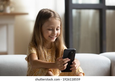 Smart Little User. Happy Small Kid Girl Sit On Sofa Alone Hold Cellphone Play Online Game. Excited Child Of Primary School Age Skilled In Using Modern Phone Gadget Watch Cute Video Having Fun At Home