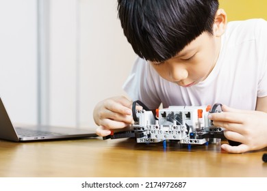 Smart Looking Asian Student Kid Assembling Robot, Coding And Solving Engineering Problem At Home With Yellow Wall In Background. STEM Education And 21st Century Learning Skills Concept