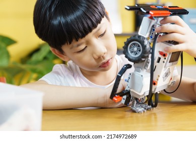 Smart Looking Asian Student Kid Assembling Robot, Coding And Solving Engineering Problem At Home With Yellow Wall In Background. STEM Education And 21st Century Learning Skills Concept