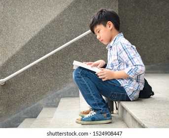 A Smart Looking Asian Tween / Teen Student Boy Sitting On The Stairs In Front Of A Library Or Collage Campus Concentrate On Reading Textbook Preparing For His Exams. Teenager Self Esteem Concept.