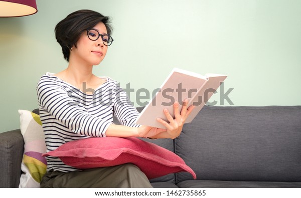 Smart looking beautiful asian middle aged\
woman 40s wearing eyeglasses reading book on the couch in living\
room. Middle aged health/beauty, Long-sightedness, Progressive\
lens. Eyes problem,\
Hyperopia.