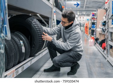 Smart Male Customer Choosing New Tires In The Supermarket For Buying. Big Shopping Mall With Car Goods.