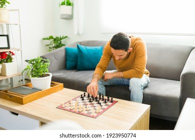 Smart Man Moving A Chess Piece While Sitting On The Sofa. Hispanic Man Playing Chess Alone As A Part Of His Mental Health Activities 