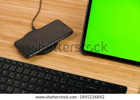 Smart phone is charging on wireless charging next to the tablet with chromo key and keyboard.