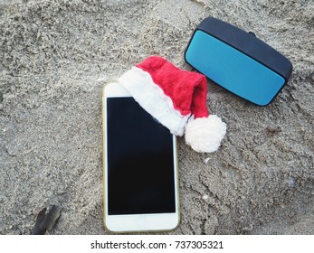 Smart Phone With Christmas Hat And Bluetooth Speaker On Beach