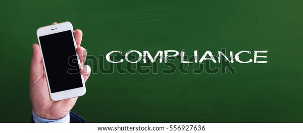 Smart phone in hand front of blackboard and\
written COMPLIANCE