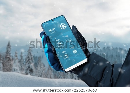 Smart phone in hand with glove with weather app on display. Winter, snowing time. Online service with weather forecast for several days concept
