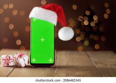 Smart Phone Mock Up With Empty Green Screen In Santa Hat On Wooden Table With Gift Boxes. Layout For Advertising Products And Services In Mobile Application. Christmas Lights In Background.