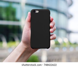 Smart Phone On A Blurry City Background In A Black Plastic Case Back View. Smart Phone In Man's Hand. Template Of Phone Case