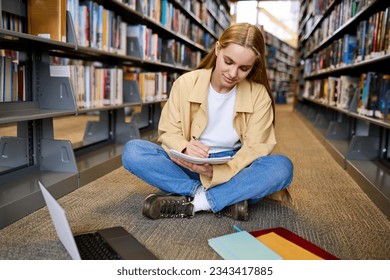 Smart pretty girl student using laptop computer sitting on floor among bookshelves in university campus library elearning, doing college course study, doing research, hybrid learning, taking notes.