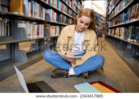 Smart pretty girl student using laptop computer sitting on floor among bookshelves in university campus library elearning, doing college course study, doing research, hybrid learning, taking notes.