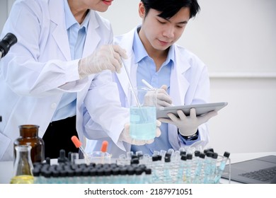 A Smart And Professional Asian Male Scientist Or Medical Technician Works With His Senior Female Supervisor In The Lab And Records An Experiment Result On His Clipboard.