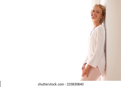 Smart professional business woman smiling and leaning on a wall by a glass door with sunny light, relaxing and enjoying her aspirational lifestyle, indoors. Home interior with businesswoman standing.