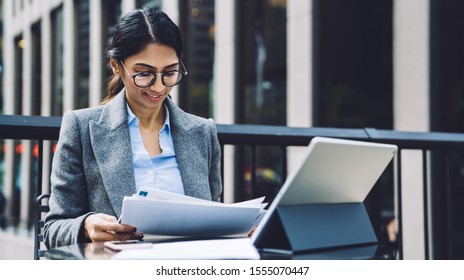 Smart professional ethnic businesswoman in elegant outfit and eyeglasses sitting at table outdoors checking documents while browsing tablet at terrace of New York city