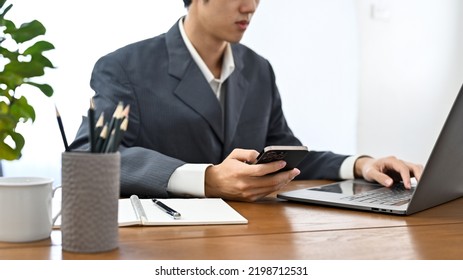 Smart And Professional Young Asian Male Banker In Formal Suit Working At His Office Desk, Using Smartphone And Laptop Computer. Cropped Image