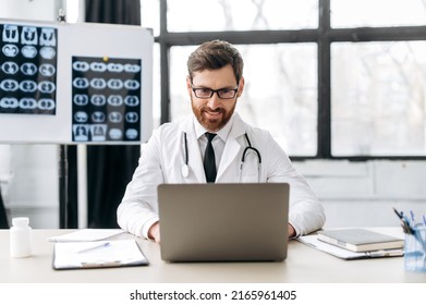 Smart proud positive caucasian male doctor of general medicine, dressed in a medical uniform and with glasses, sits at his workplace in a medical office, looking in a laptop, consults patients online