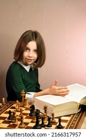 Smart And Purposeful Young Female Chess Player Reflects On Position Of Pieces In Chess Game. Schoolgirl With Huge Book Analyzes And Studies Position On Chessboard. Chess Treatise