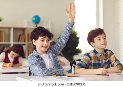Smart schoolboy sits at a desk next to his classmate in class with his hand raised, wanting to give the right answer or participate. Education, primary school, learning and people concept.
