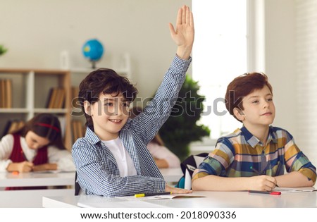 Smart schoolboy sits at a desk next to his classmate in class with his hand raised, wanting to give the right answer or participate. Education, primary school, learning and people concept.