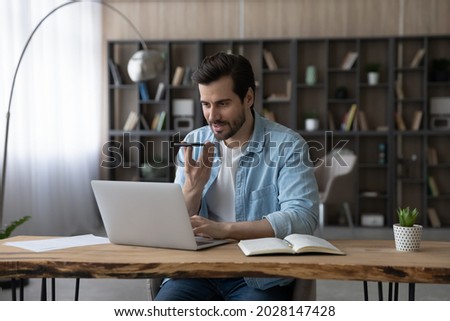Smart skilled happy young businessman sitting at table with computer in modern home office, recording audio message for client on smartphone or holding voicemail distant conversation with colleagues.