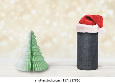 Smart Speaker Device Next To A Paper Christmas Tree On A Wooden Table. Light Bokeh Background And Empty Copy Space For Editor's Text.