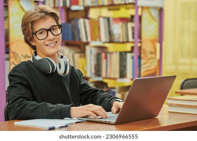 A smart student in glasses and headphones looks happily at the camera while sitting at a table in a library and studying with a laptop. Education. Modern students. - Powered by Shutterstock