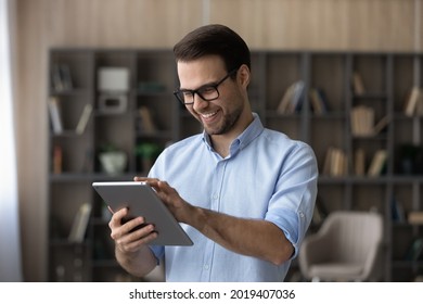Smart tech. Positive guy in spectacles use touch pad device to learn or work online choose goods services at web shop scroll news at social media. Young man stand at living room play game on tablet pc