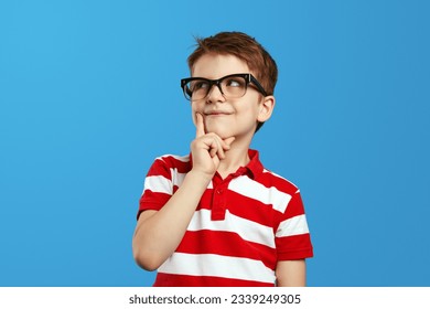 Smart thoughtful preschool boy in nerdy glasses and red striped polo shirt touching cheek and looking up while thinking against blue background. - Powered by Shutterstock