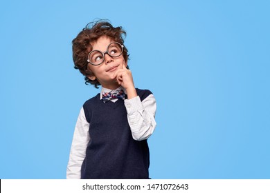 Smart wunderkind in nerdy glasses and school uniform touching cheek and looking up while thinking against blue background