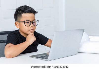 Smart Young Asian Boy Wearing Glasses And Black Shirt Sitting On Chair Of White Working Desk In Home Office, Seriously Catching Chin And Concentrate On Looking At Laptop To Consider Difficult Info