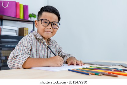 Smart young Asian boy wearing glasses and grey checked shirt sitting on studying desk of tablet, mobile phone, and colorful pencils in reading room and concentrate on drawing on white paper - Powered by Shutterstock