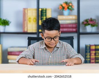 Smart young Asian boy wearing glasses and grey checked shirt happily sitting in reading room and enjoy study of searching funny info by tablet on desk