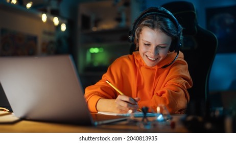 Smart Young Boy In Headphones Attending Online Class On Laptop Computer In Cozy Dark Room At Home. Happy Teenager Browsing Educational Research Online, Writing In Notebook, Studying School Homework.