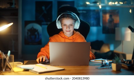 Smart Young Boy In Headphones Using Laptop Computer In Cozy Dark Room At Home. Happy Teenager Browsing Educational Research Online, Chatting With Friends, Studying School Homework.