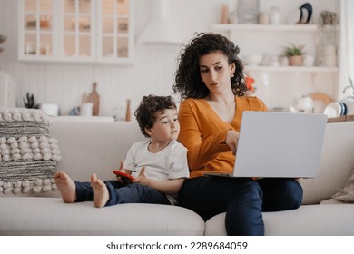 Smart young Italian woman with curly hair dressed in orange blouse and blue pants sitting on sofa using laptop, working at home at the kitchen with son playing on phone, talking with mom. Family - Powered by Shutterstock