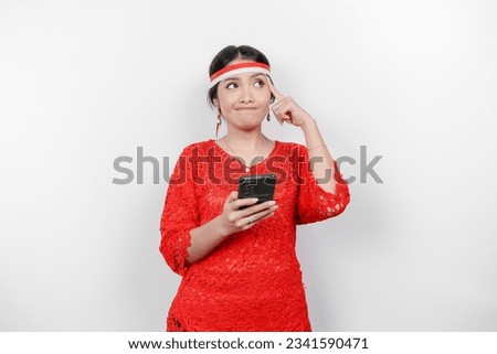 A smart young woman wearing headband and red kebaya is looking aside to an idea on copy space while holding her phone, isolated by white background. Indonesia's independence day.