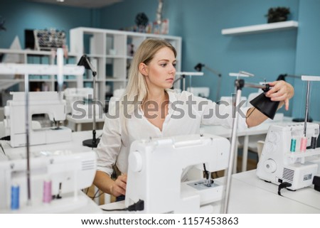 A smart-looking pretty blond woman wearing white shirt is sewing with the electric sewing-machine. Fashion, tailor's workshop.