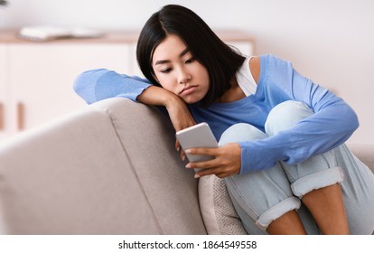 Smartphone Addiction And Depression. Portrait of frustrated bored asian young lady using mobile phone, checking social media, reading bad news sitting on the couch. Upset woman waiting for his call