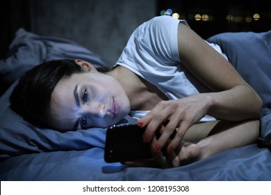 Smartphone Addiction. Young Tired Female Looking At Her Mobile Phone Screen, Lying In Bed Late At Night, Scrolling Her Social Media News Feed
