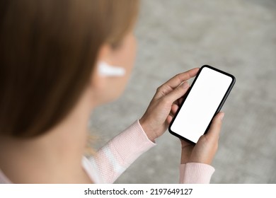 Smartphone With Blank Empty Screen Copy Space In Female Hands Close Up. Woman With White Wireless Earbud In Ear Using Mobile Phone For Listening To Music, Video Call. Online Communication