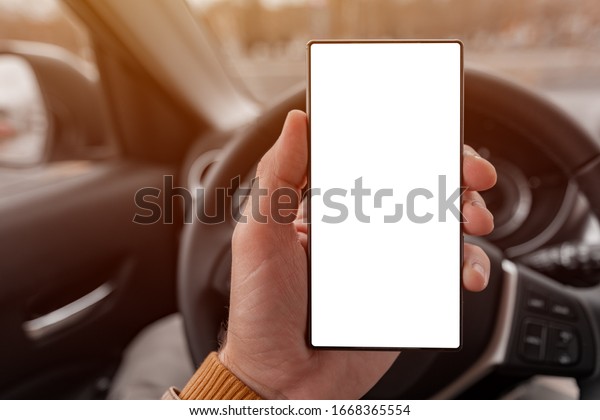 Smartphone blank screen mock up in\
driver\'s hand in front of the car steering wheel, selective\
focus