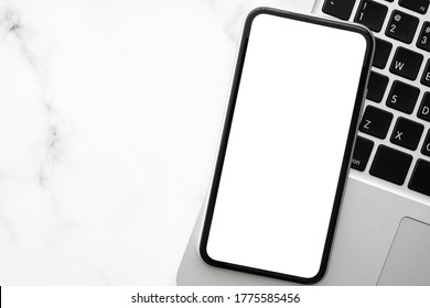 Smartphone with blank screen is on top of white marble office desk table with laptop computer. Top view with copy space, flat lay.