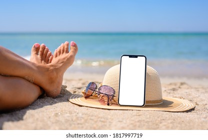 Smartphone With Blank White Display On The Beach. Hat, Sunglasses And Legs Of The Girl On The Background Of The Sea. The Concept Of Using Gadgets And The Internet At A Seaside Resort.