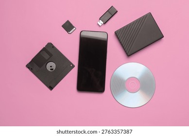 Smartphone with CD disk, floppy disk, USB flash drive, sd card and external hard drive on pink background. Digital information media. Flat lay