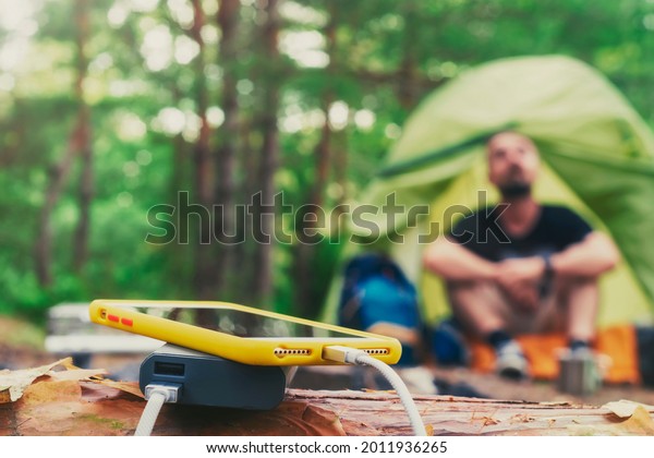 Smartphone is charged using a portable charger.\
Power Bank charges the phone outdoors with a backpack against the\
backdrop of a tent and a\
tourist